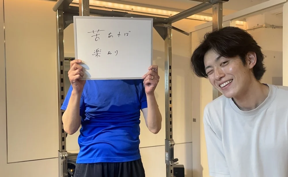WORKOUTHOLIC下北沢店のトレーニングルームで「苦あれば楽あり」と書いたホワイトボードを掲げる60代男性会員と、隣で笑顔のトレーナーの写真