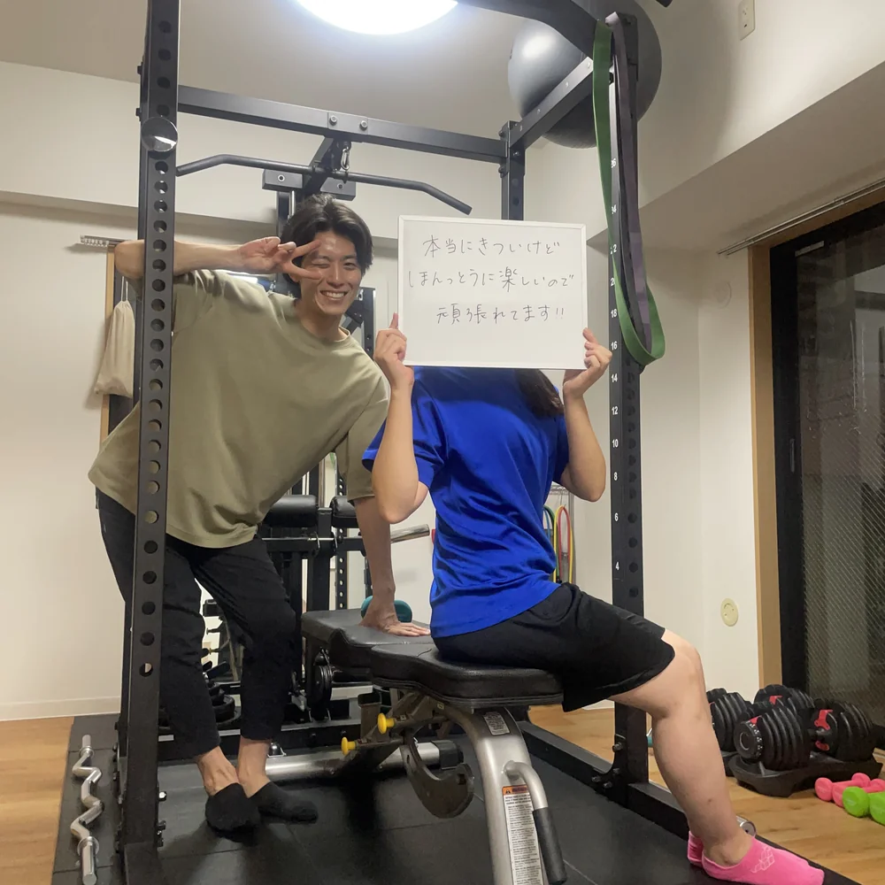 WORKOUTHOLIC横浜店でピースサインをするトレーナーと、「本当にきついけどほんっとうに楽しいので頑張れてます!!」と書いたホワイトボードを掲げる20代女性会員の写真
