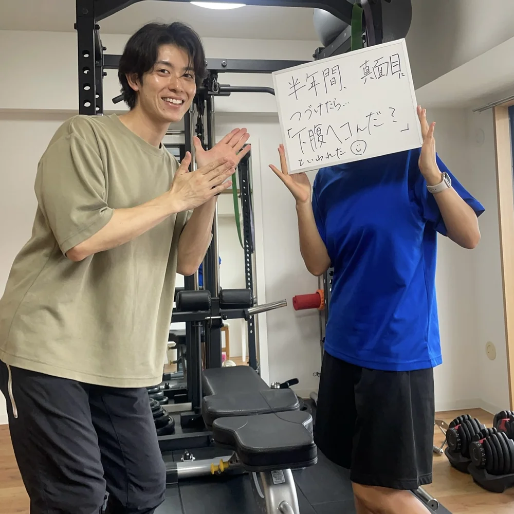 WORKOUTHOLIC横浜店で「半年間、真面目につづけたら…下腹へこんだ？といわれた」と書いたホワイトボードを掲げる30代女性会員と、隣で両手を広げるトレーナーの写真